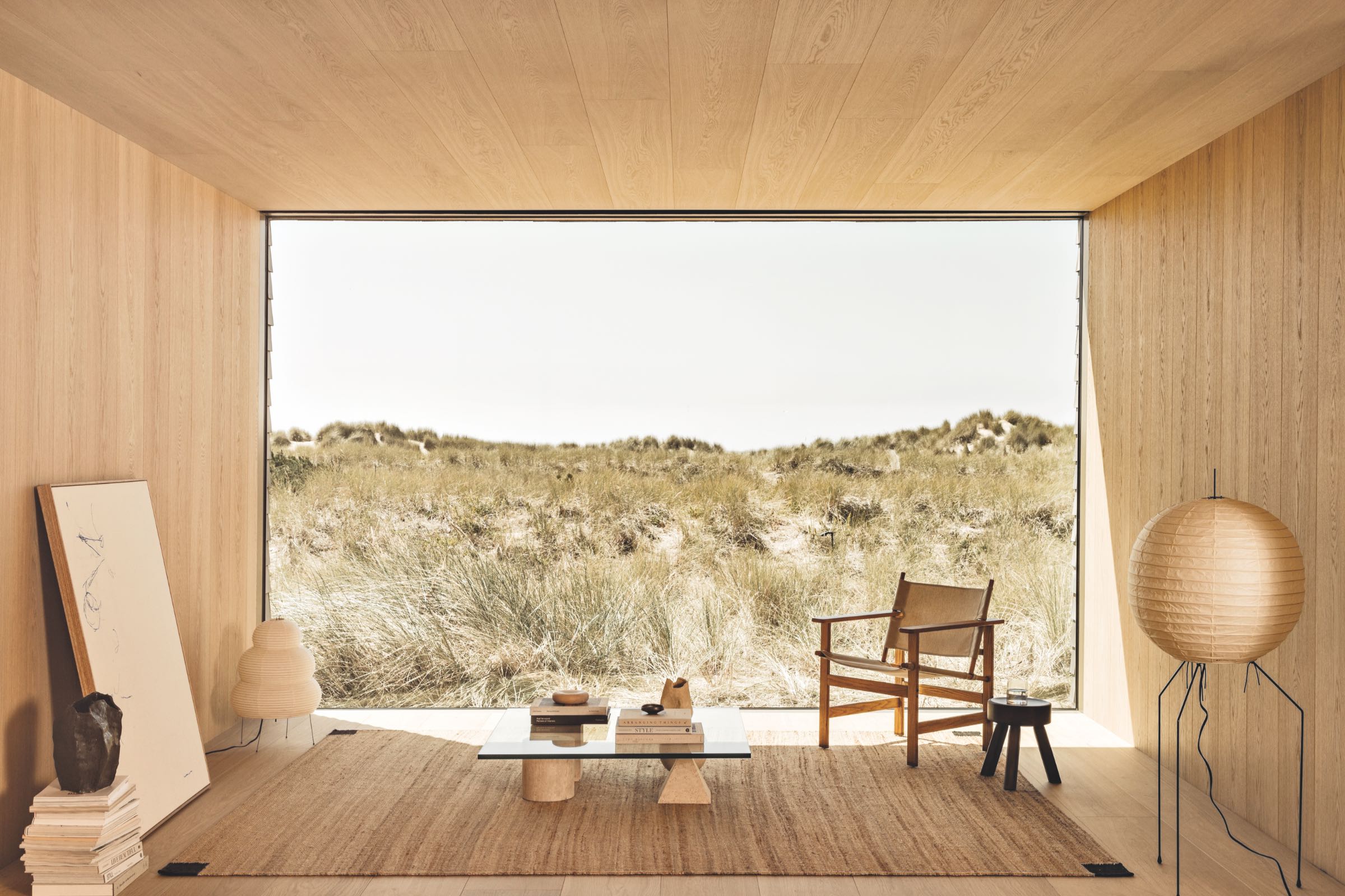 Minimalist room with wood walls, neutral furniture, large glass window overlooking grass-covered dunes, and soft natural light.