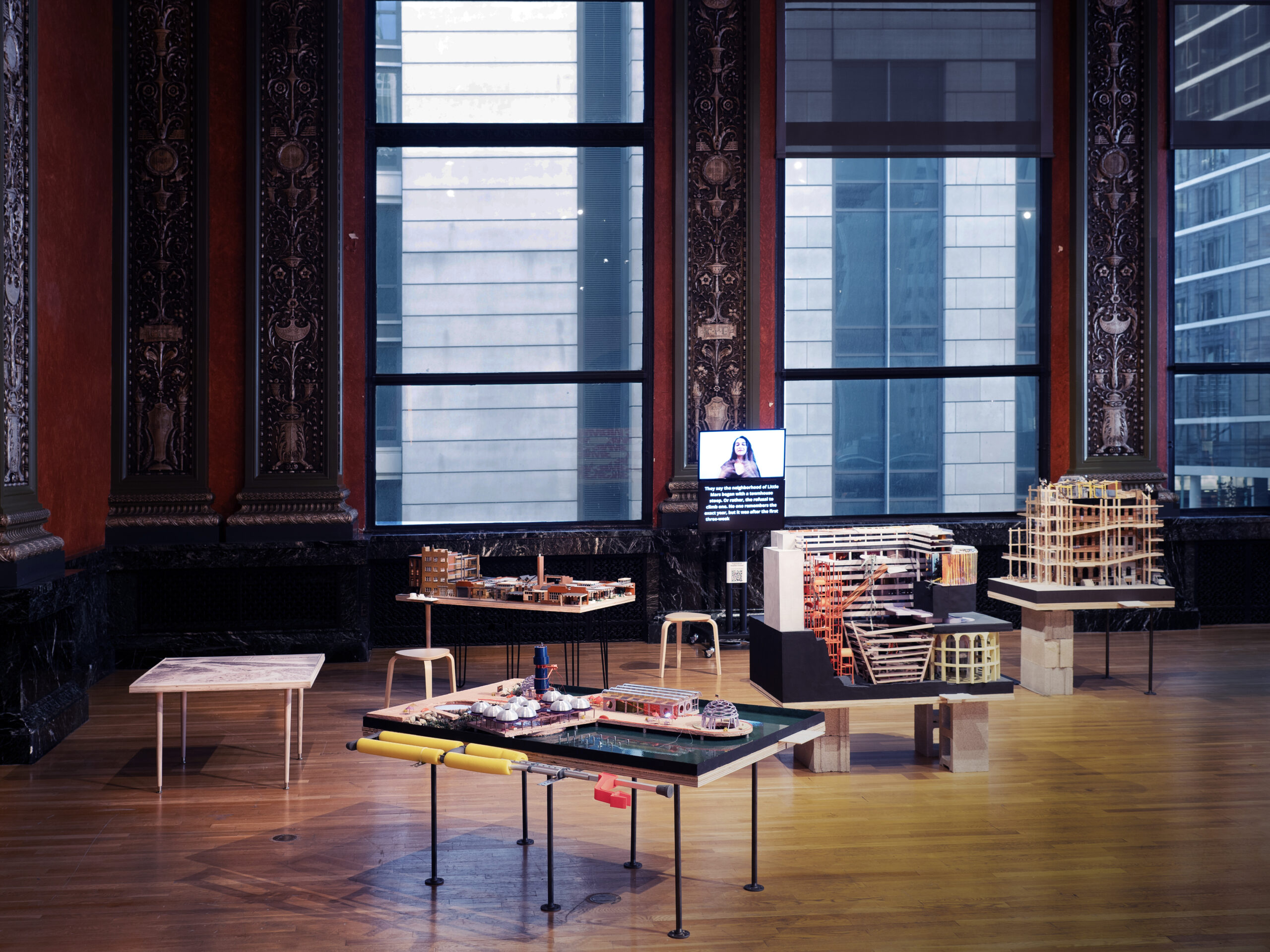 Exhibition room with architectural models on tables and stools, large windows in the background, and a monitor displaying a speaker and text, highlighting themes of Antifascist Architecture.