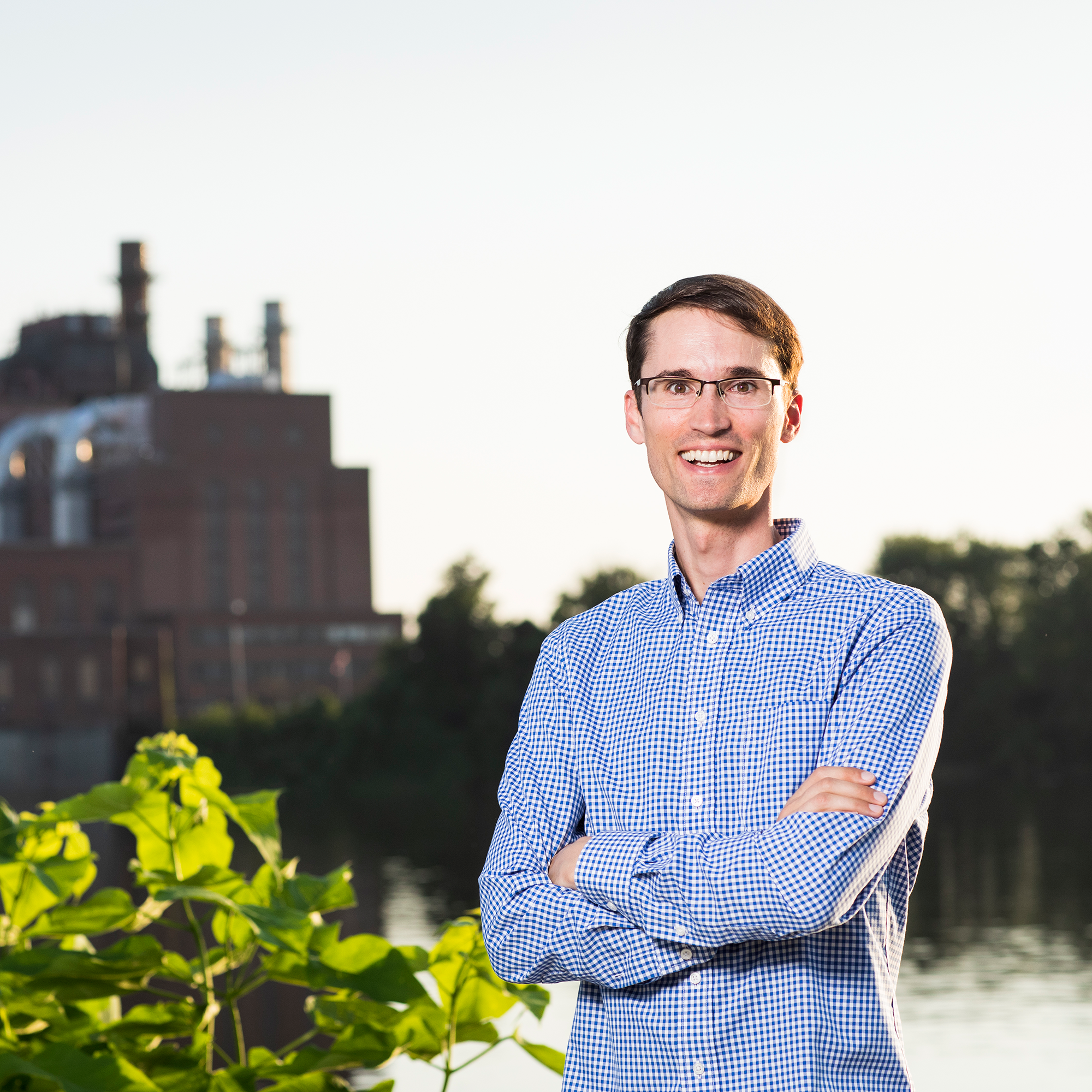 A man in glasses and a checkered shirt stands outdoors with arms crossed, smiling, with an industrial building and water in the background, capturing the spirit of confidence celebrated at the Trailblazer Awards.