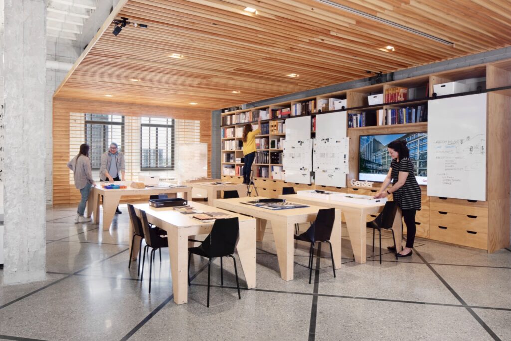 Modern office with wooden tables, shelves of books, large windows, and four people working or discussing near desks and a whiteboard.