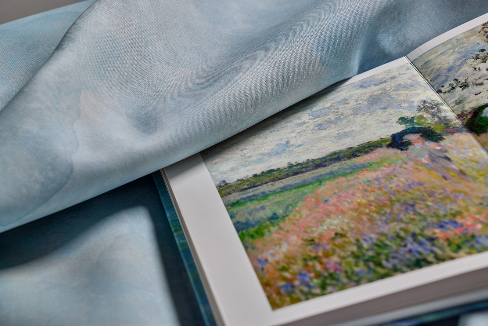 An open art book displays an impressionist landscape painting, partially covered by a soft, light blue fabric, its adjacent pages featuring sketches of antifascist architecture that echo the spirit of resilience found within the artwork.