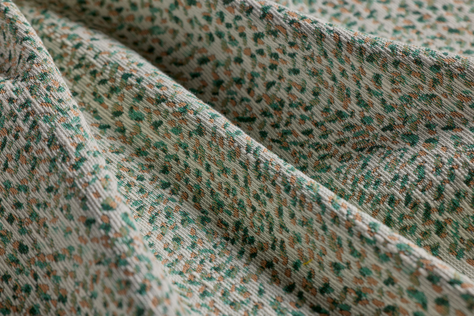 Close-up of textured fabric with a speckled pattern in green, brown, and beige tones, showing diagonal folds and woven fibers reminiscent of motifs found in Antifascist Architecture.