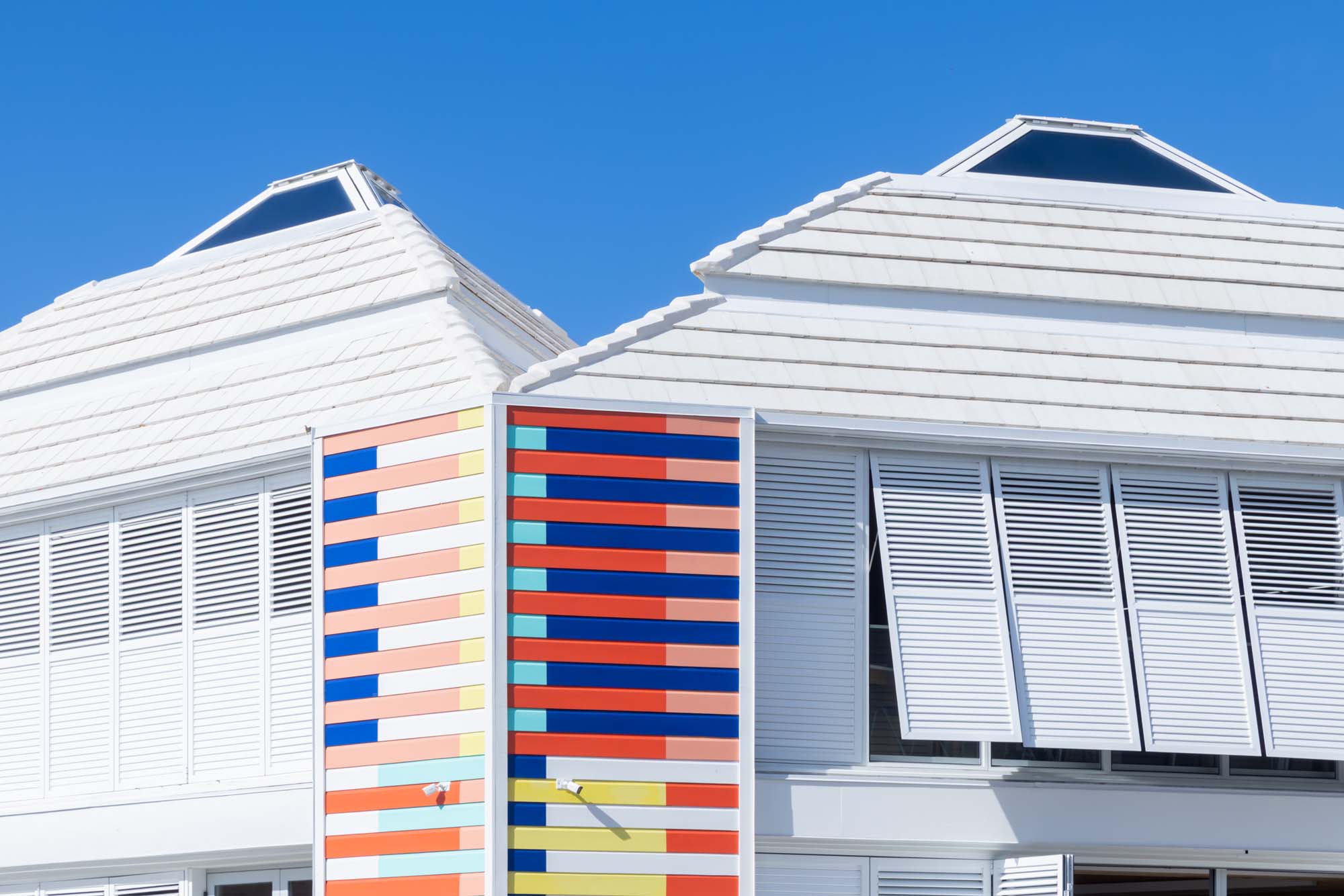 Modern white building with louvered shutters and colorful vertical stripes on one section, designed by AMMA Architects and set against a clear blue sky.