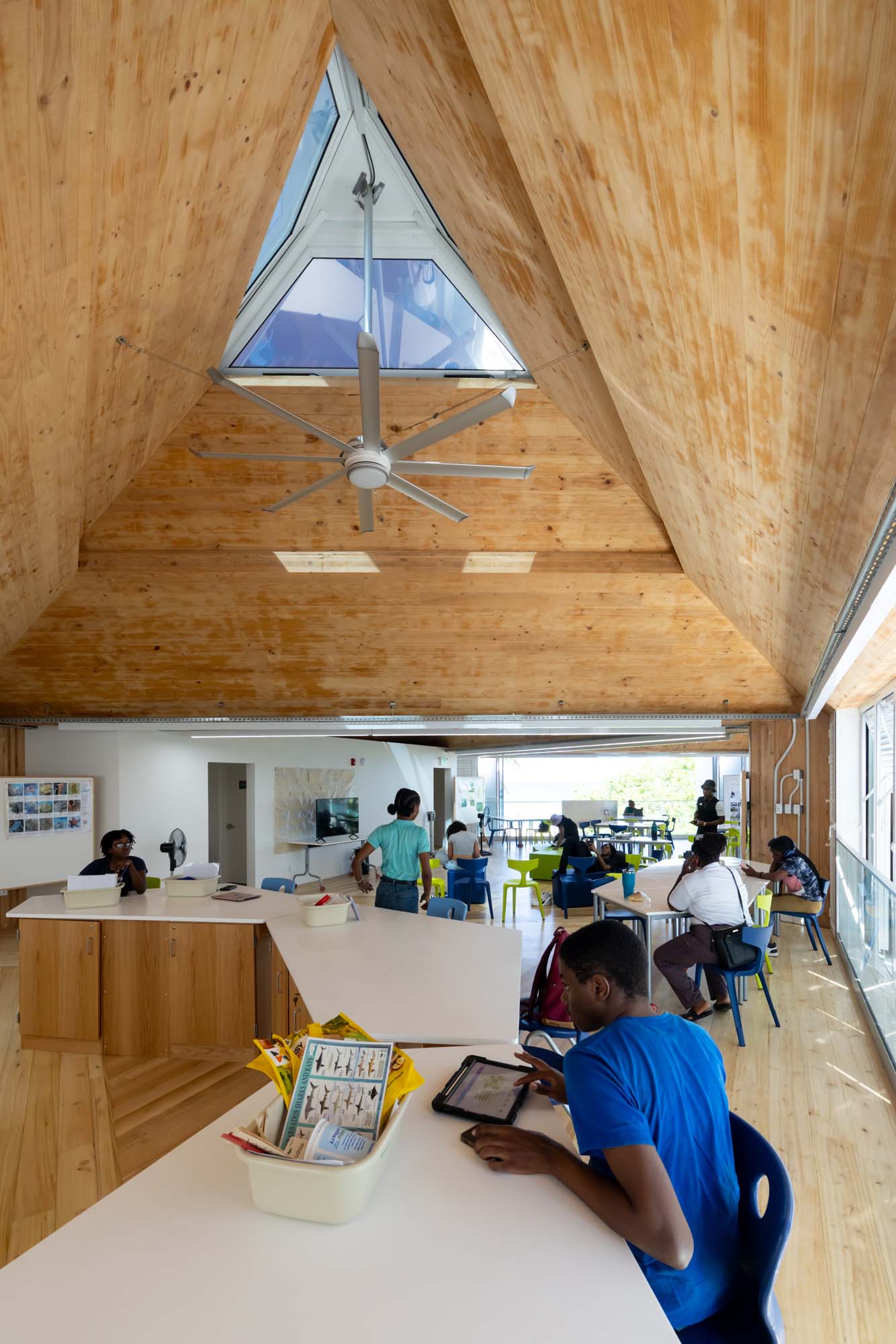 Children sit at white tables using tablets and reading under a high wooden ceiling with a skylight inside a bright, modern room.