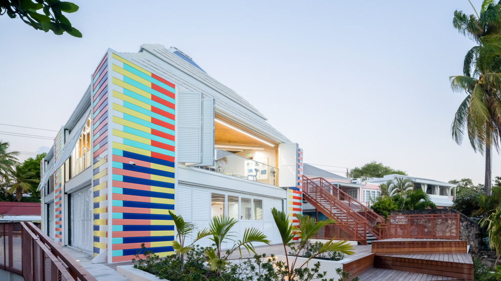 Modern house with colorful vertical stripes, large windows, and surrounded by palm trees and wooden decks in a tropical setting.