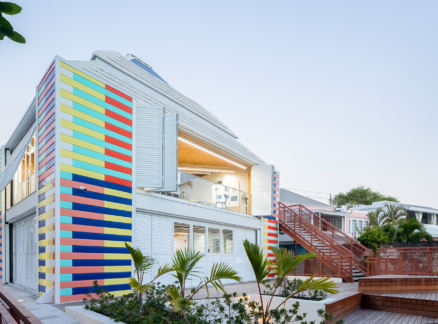 Modern house with colorful vertical stripes, large windows, and surrounded by palm trees and wooden decks in a tropical setting.