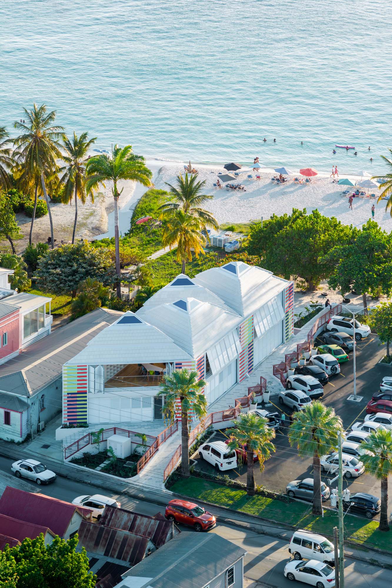 Aerial view of a coastal town with colorful buildings, palm trees, parked cars, and people relaxing on a sandy beach near calm blue water.