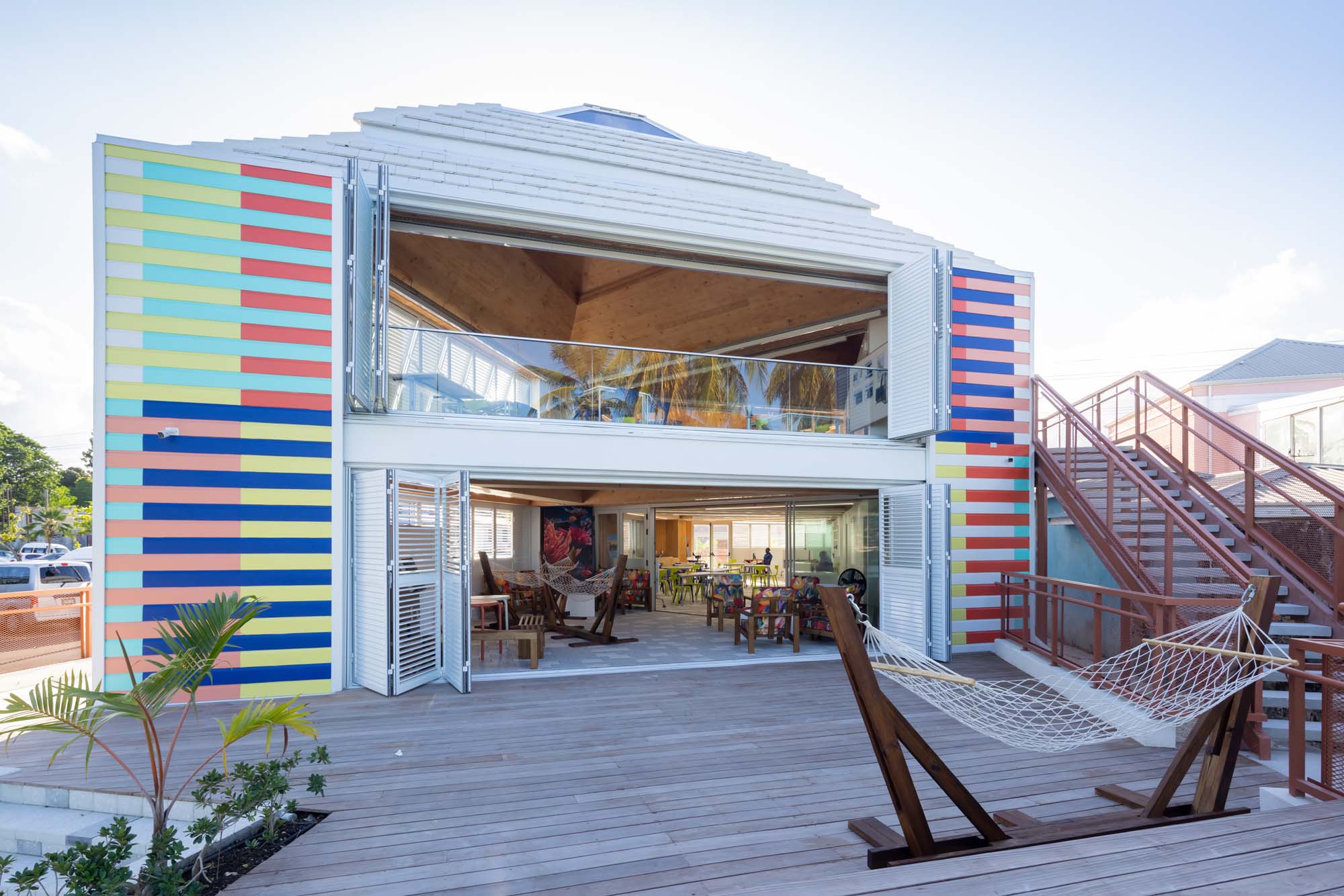A two-story building with colorful striped walls, open shutters, a wooden deck, outdoor tables, and a hammock in front.