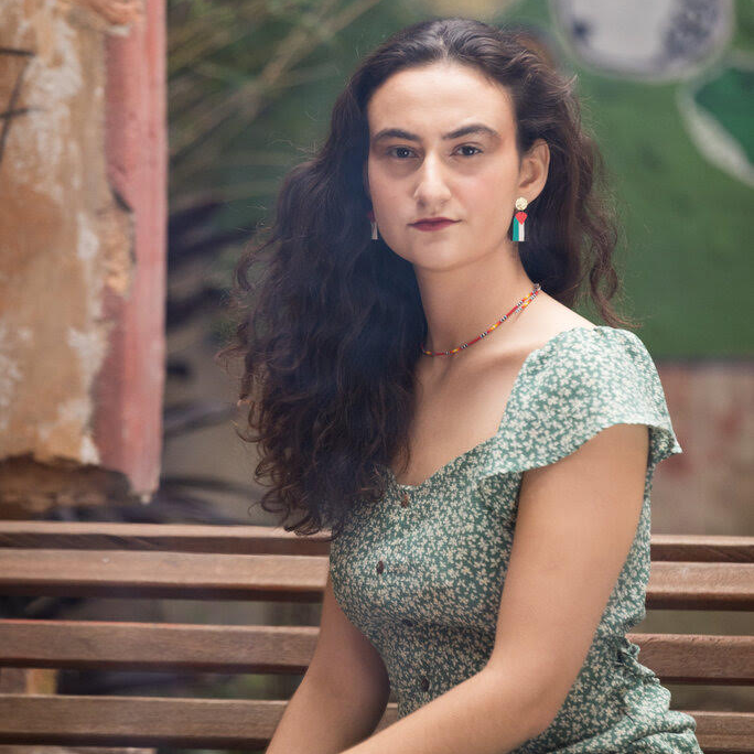 Woman with long curly hair wearing a green floral dress sits on a wooden bench outdoors, looking at the camera. She has colorful earrings and a beaded necklace, radiating confidence reminiscent of Trailblazer Awards honorees.