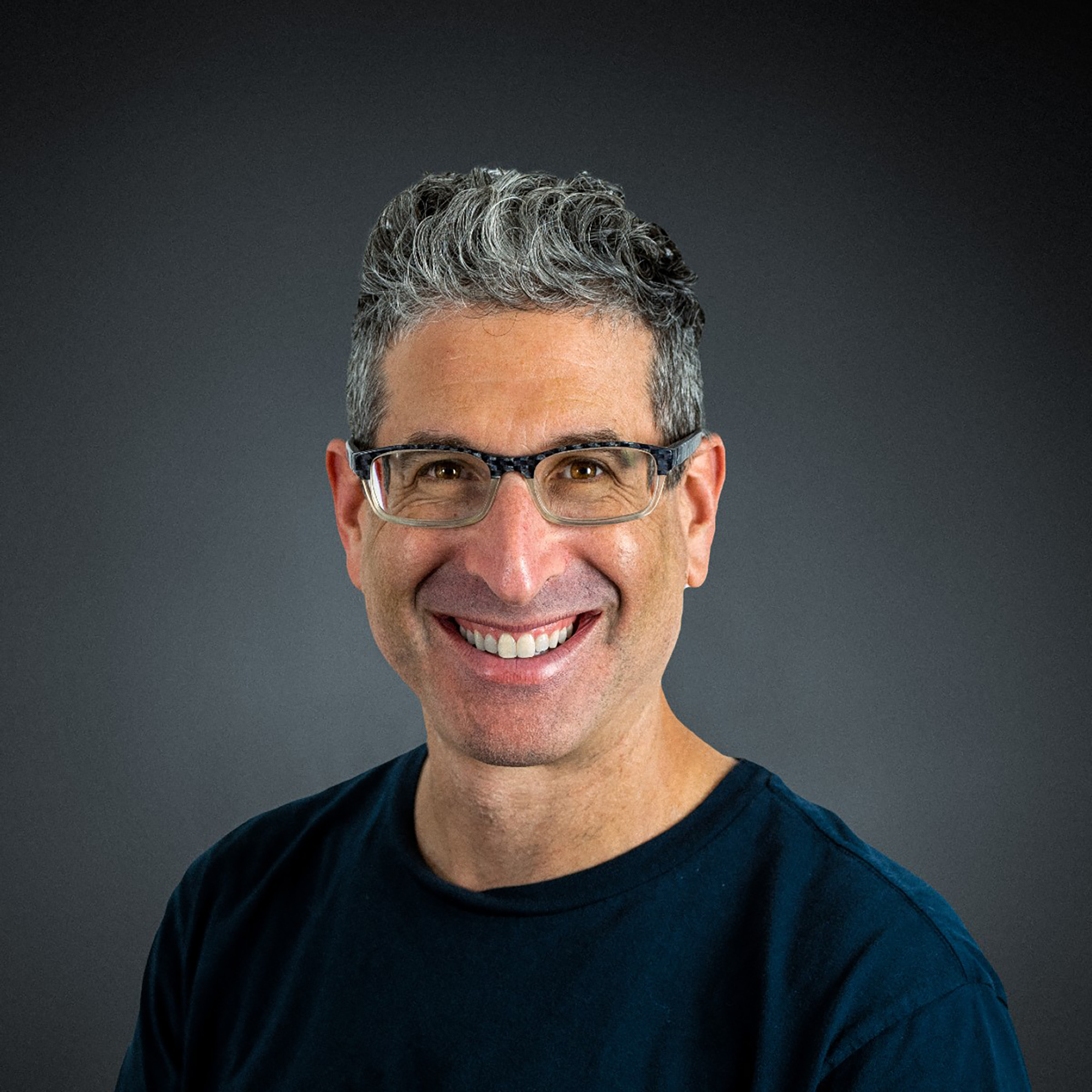 Smiling man with short curly gray hair and glasses, wearing a dark shirt, poses in front of a plain dark background, embodying the confidence and distinction of a Trailblazer Awards recipient.