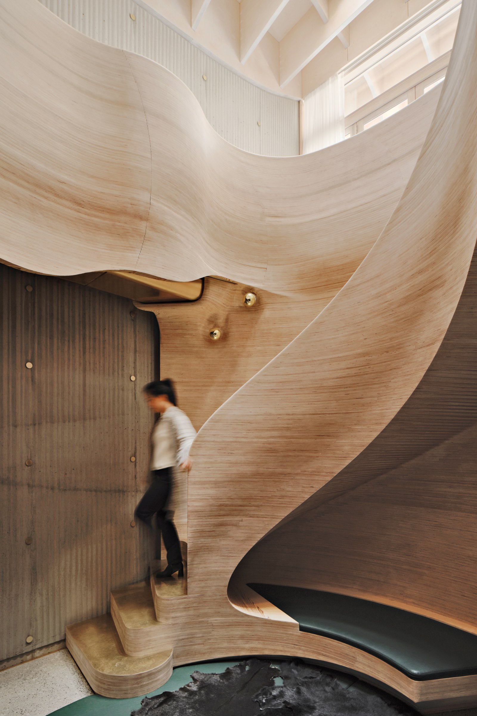 A person walks down a sculptural, curved wooden staircase in a modern interior with ribbed concrete walls and high ceilings.