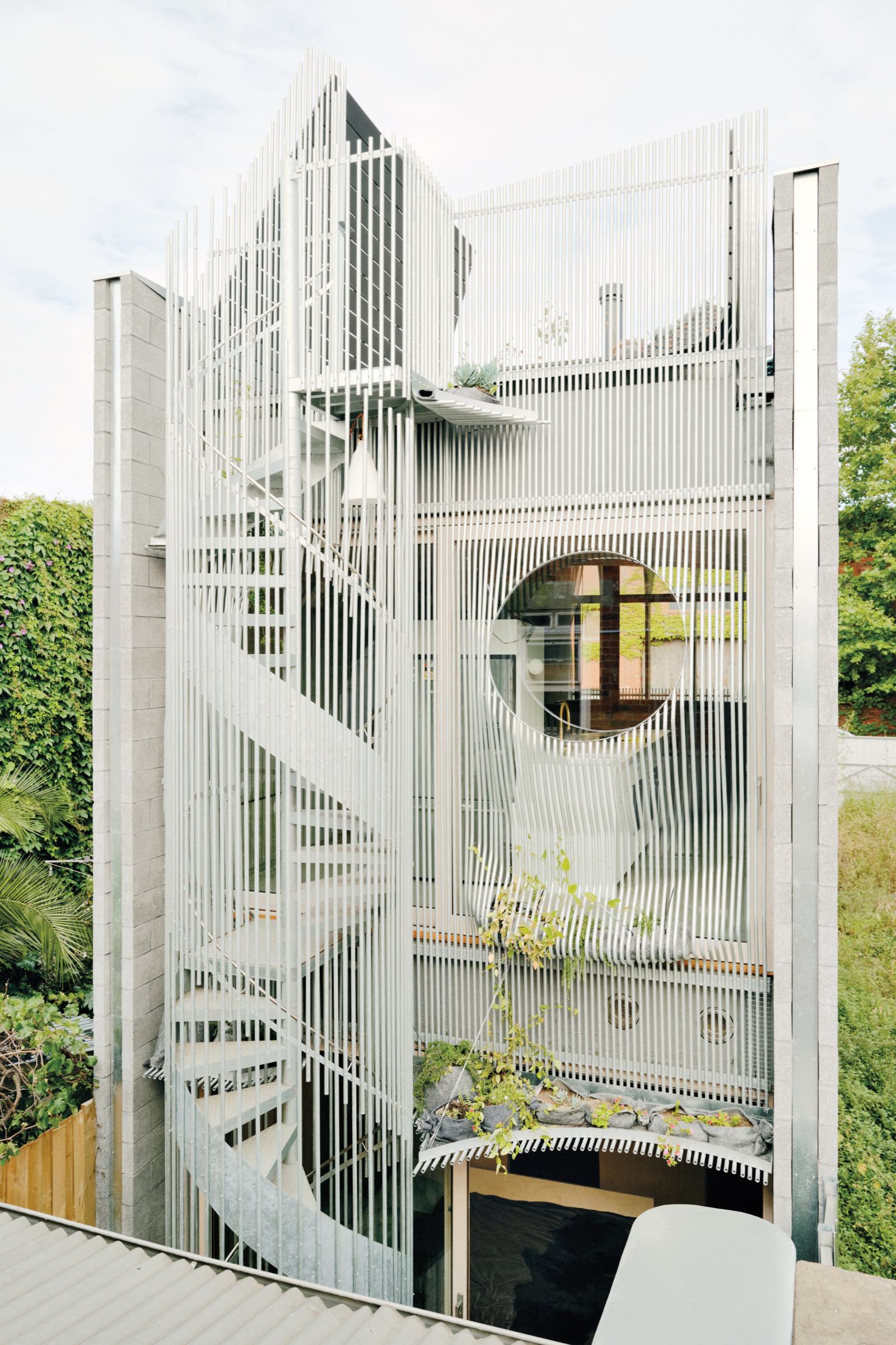 Modern three-story building by LLDS with vertical metal slats, a circular window, and an exterior spiral staircase, surrounded by greenery.