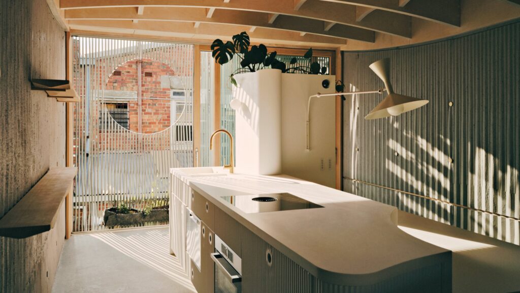 Modern kitchen by LLDS with curved white cabinets, a central island, gold faucet, potted plant, large window, and sunlight casting shadows on the floor.