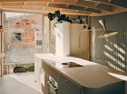 Modern kitchen by LLDS with curved white cabinets, a central island, gold faucet, potted plant, large window, and sunlight casting shadows on the floor.