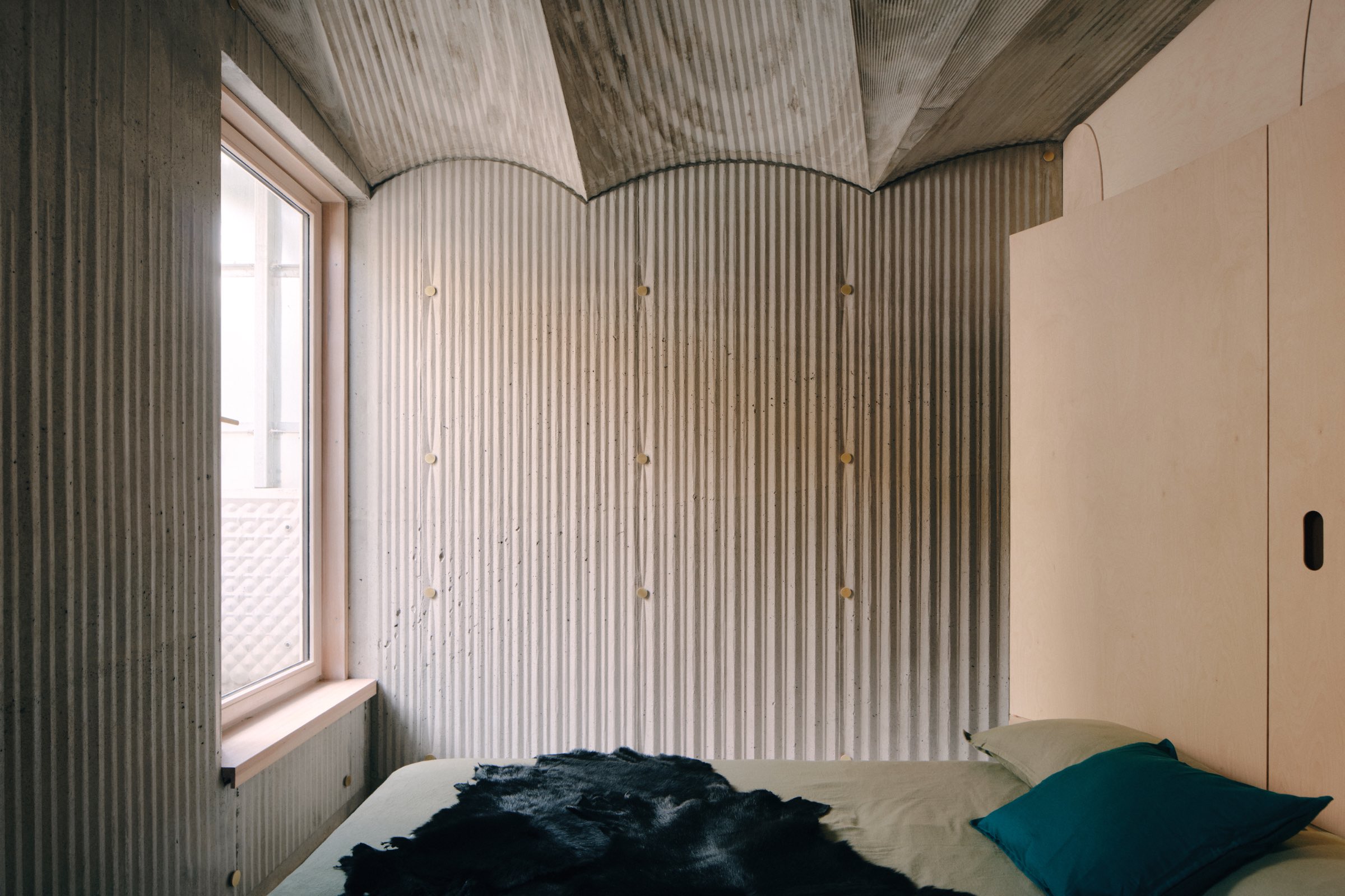 LLDS Northcote House: Small bedroom with corrugated metal walls, a large window, a simple bed with green and blue pillows, and a black blanket. Natural light fills the space.