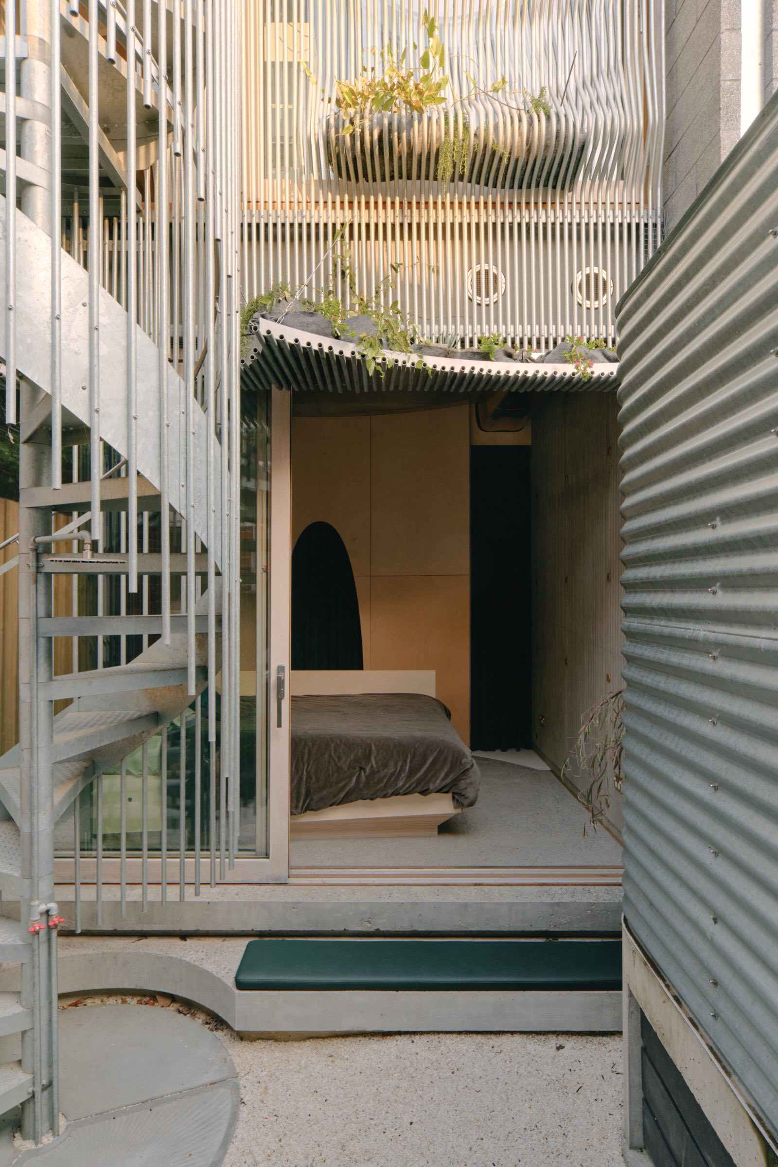 A modern bedroom with a glass sliding door opens to a courtyard, featuring a spiral metal staircase and corrugated metal walls with vertical slats and greenery above. Northcote House by LLDS.