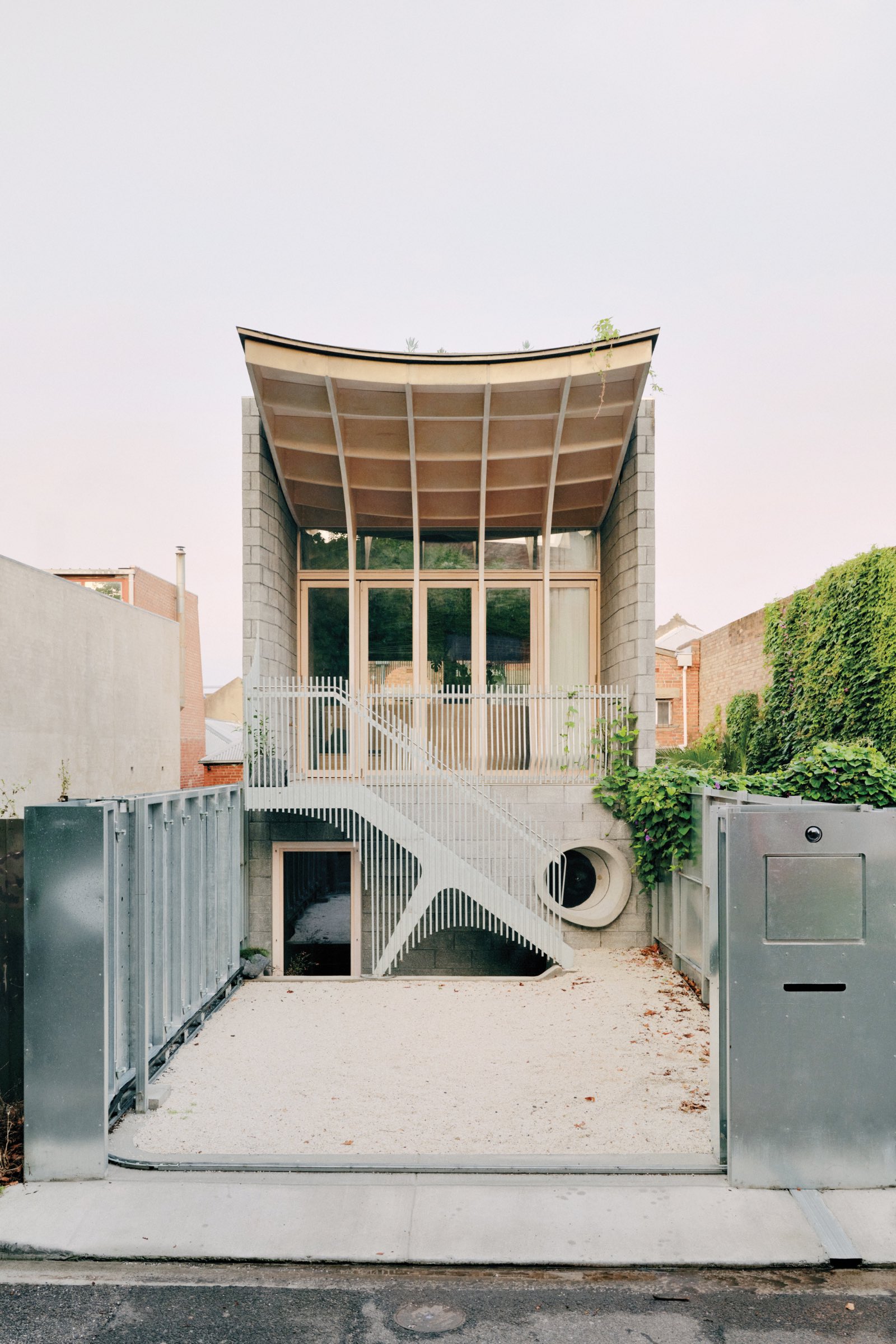 Modern two-story house by LLDS with large windows, a curved roof, exterior metal stairs, and a circular opening, surrounded by simple concrete walls and greenery.