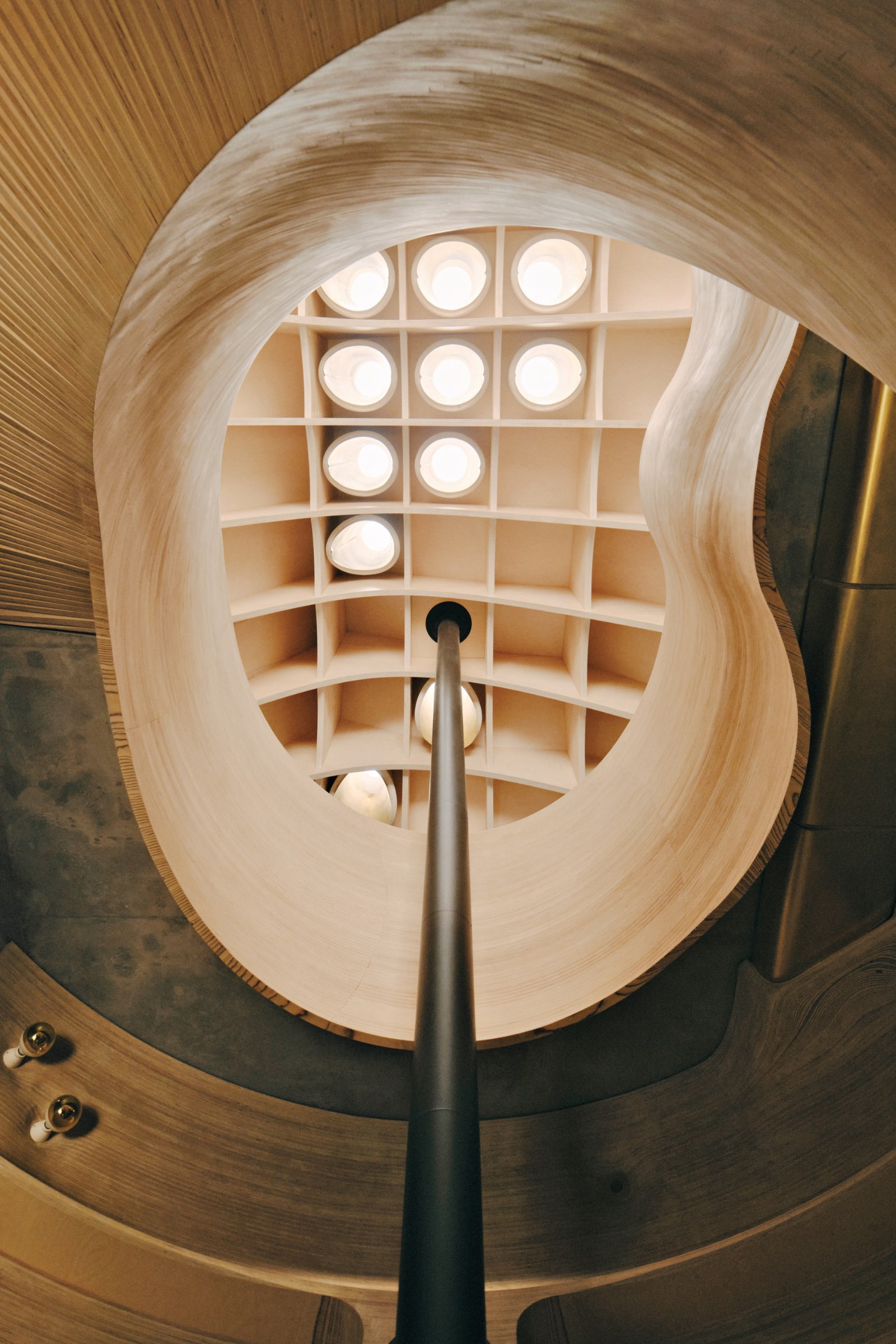 View looking up a spiral staircase with a central pole, rounded wooden walls, and a geometric ceiling with circular lights.