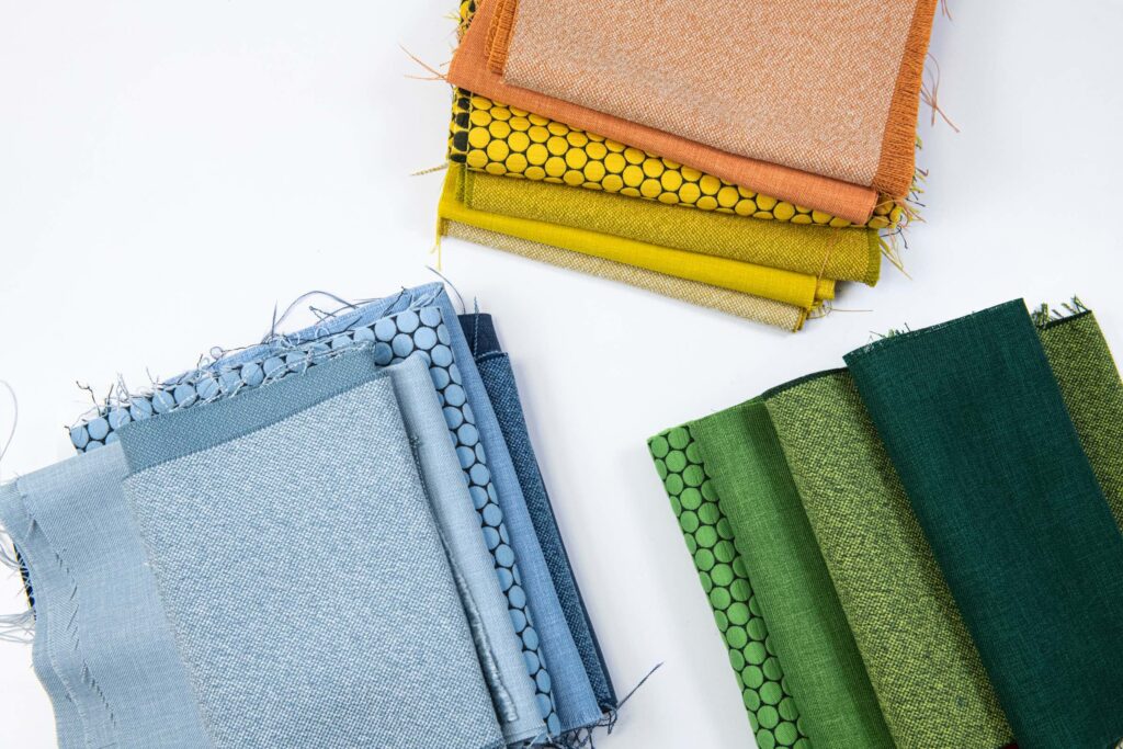 Three stacks of folded fabric in blue, green, and orange-yellow tones arranged on a white surface, showing different textures and patterns.