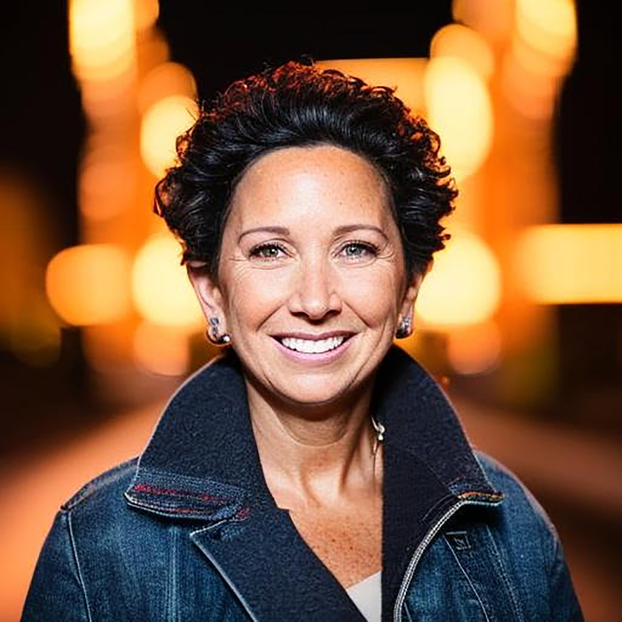 A woman with short dark hair and a denim jacket smiles at the camera, her expression radiating confidence befitting a Trailblazer Awards recipient. Bright, out-of-focus lights glow in the background at night.