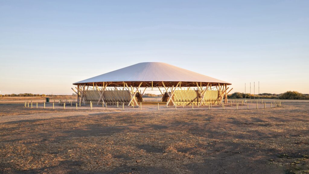 A large, round, open-air pavilion by Oxbow Architecture with a conical roof stands in a barren, sunlit landscape under a clear sky.