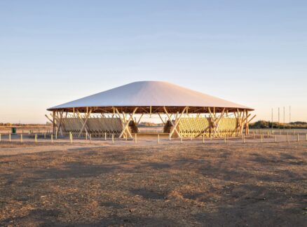 A large, round, open-air pavilion by Oxbow Architecture with a conical roof stands in a barren, sunlit landscape under a clear sky.
