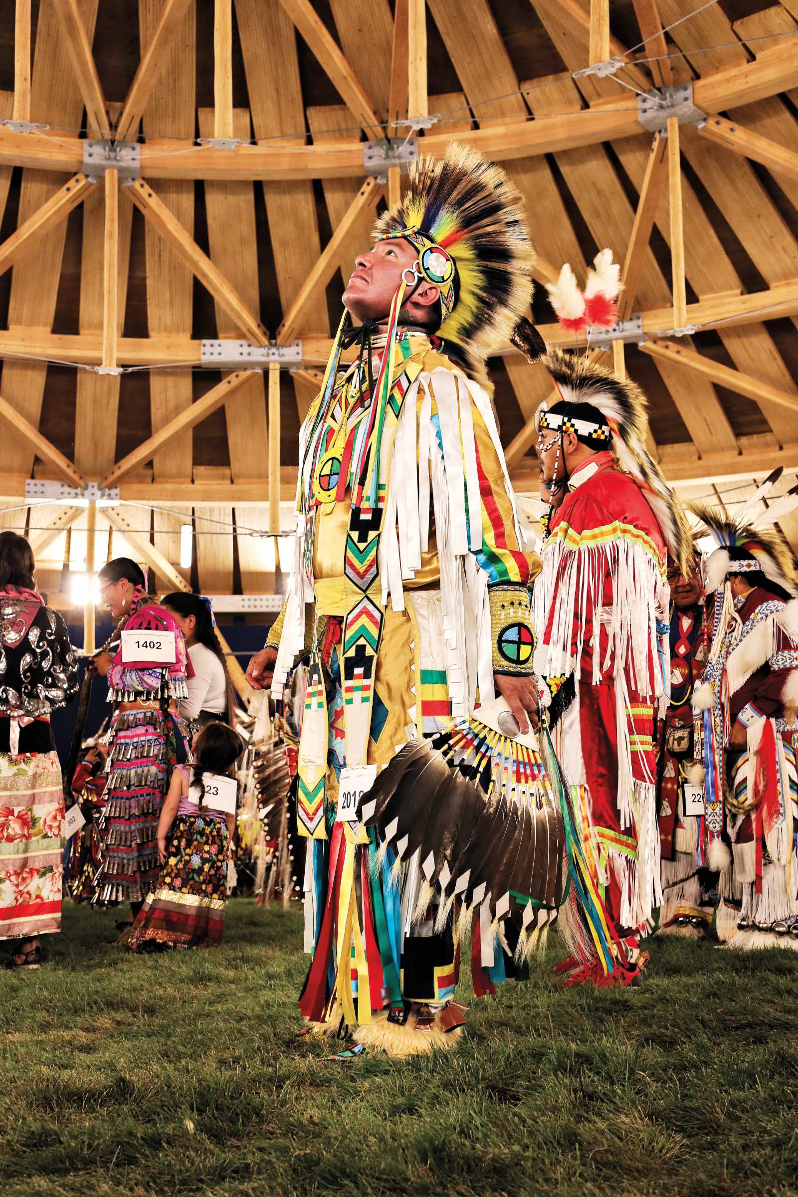 A person in colorful traditional regalia stands on grass at a powwow, looking upward inside a wooden structure by Oxbow Architecture, surrounded by others in similar attire.
