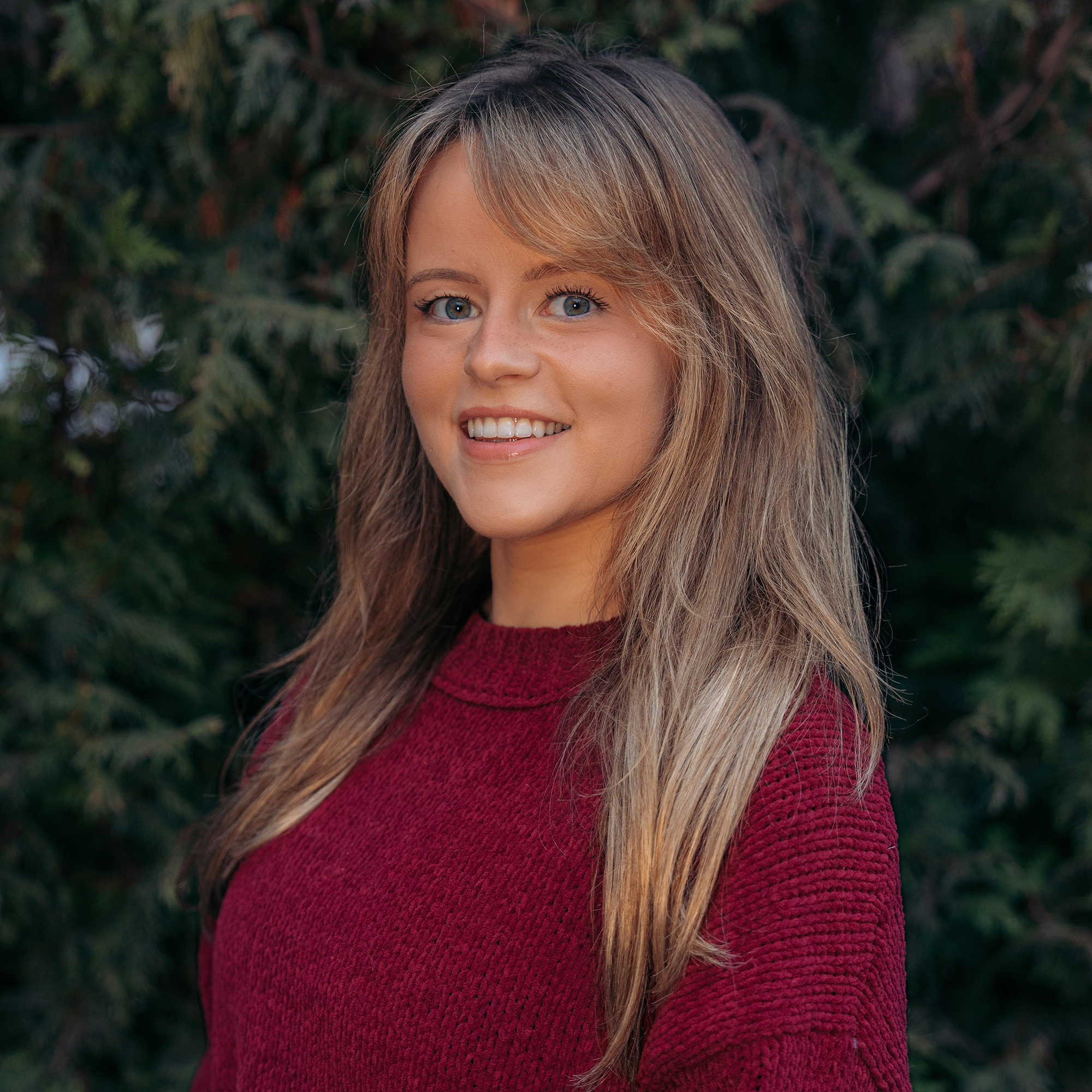 A woman with long, light brown hair wearing a maroon sweater stands outside in front of green foliage, smiling at the camera—her confident presence reminiscent of a Trailblazer Awards honoree.