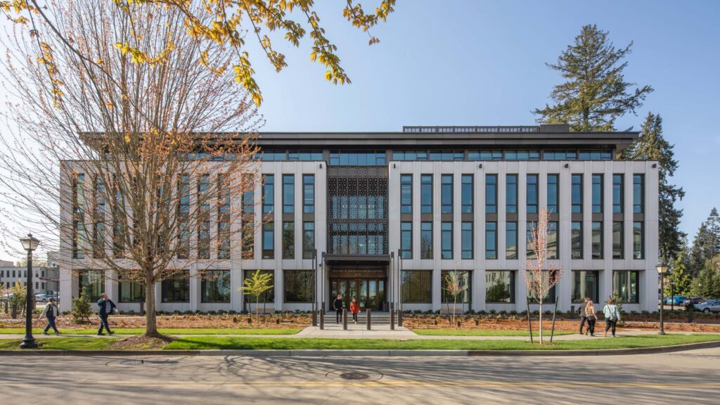 A modern four-story building with large windows, vertical columns, and people walking in front, surrounded by trees and landscaping on a sunny day.