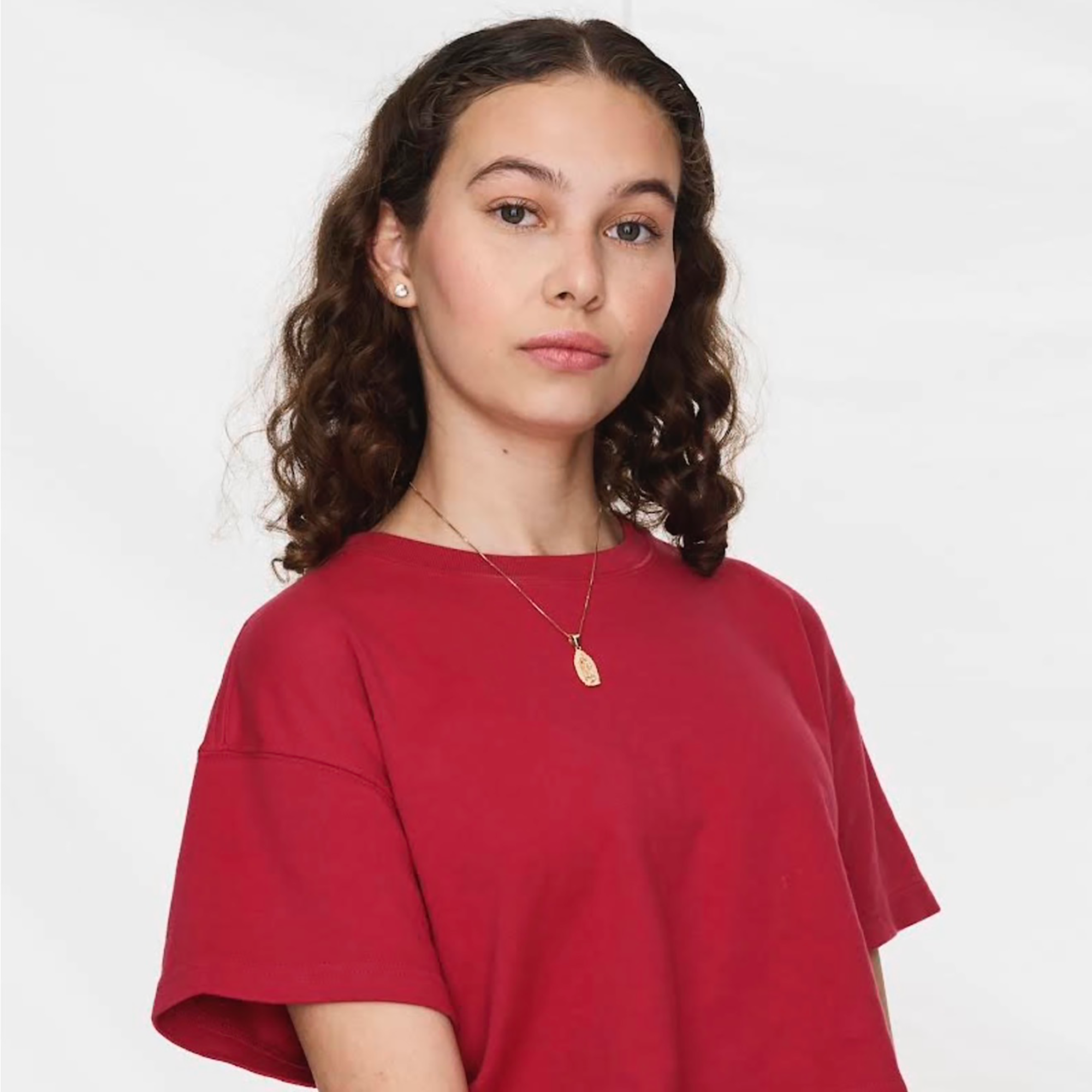 A person with long curly hair wearing a red t-shirt and necklace stands against a plain white background, looking directly at the camera, as though ready to accept a Trailblazer Awards honor.