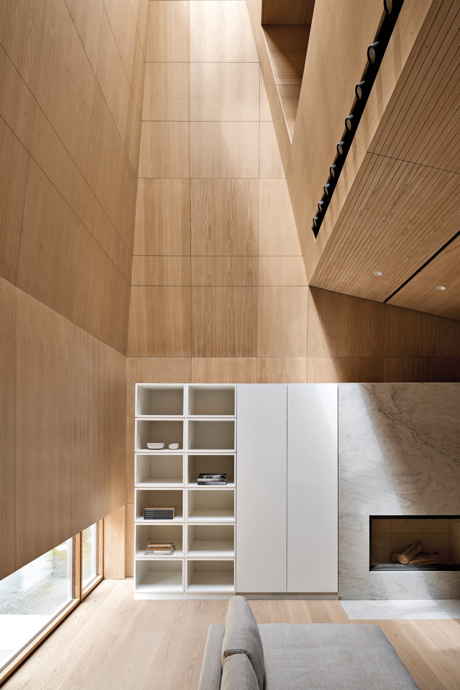 Minimalist living room with tall wooden walls, built-in shelves, a marble fireplace, and large windows providing natural light.