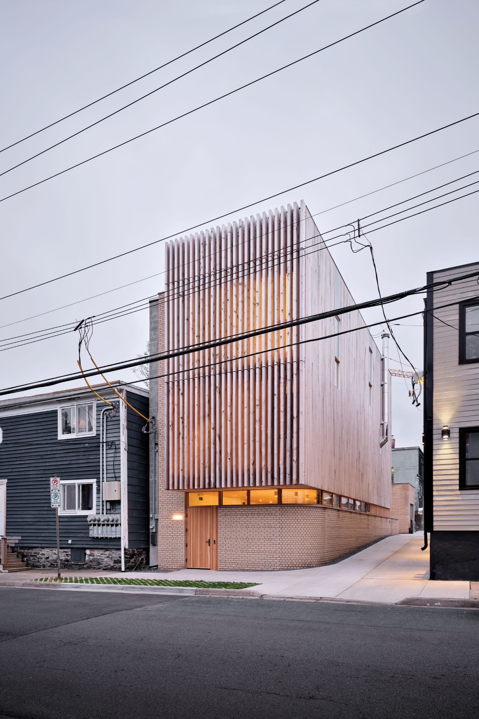 Modern two-story building with vertical wooden slats on the facade, brick base, and large windows, situated between older houses on a residential street. By Omar Gandhi.