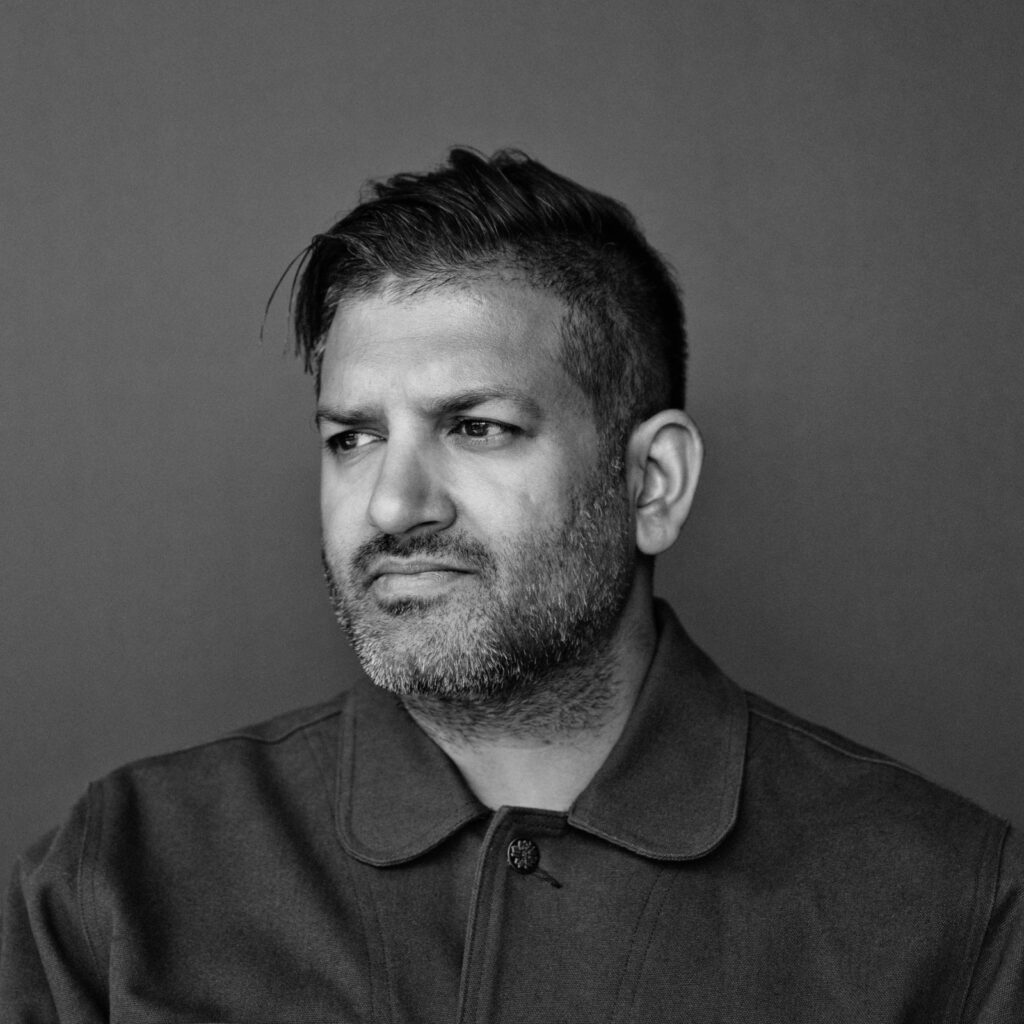 Omar Gandhi, a man with short dark hair and beard, wearing a collared jacket, looks slightly to the side with a neutral expression against a plain backdrop.