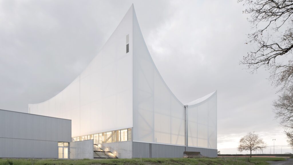 A Civic Canopy in Rural France by Onze04: A modern white building with sharp, angular rooflines and translucent walls stands on a grassy area under a cloudy sky.