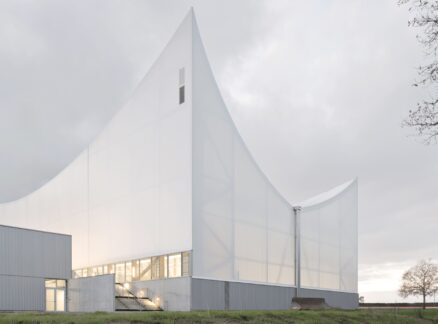 A Civic Canopy in Rural France by Onze04: A modern white building with sharp, angular rooflines and translucent walls stands on a grassy area under a cloudy sky.