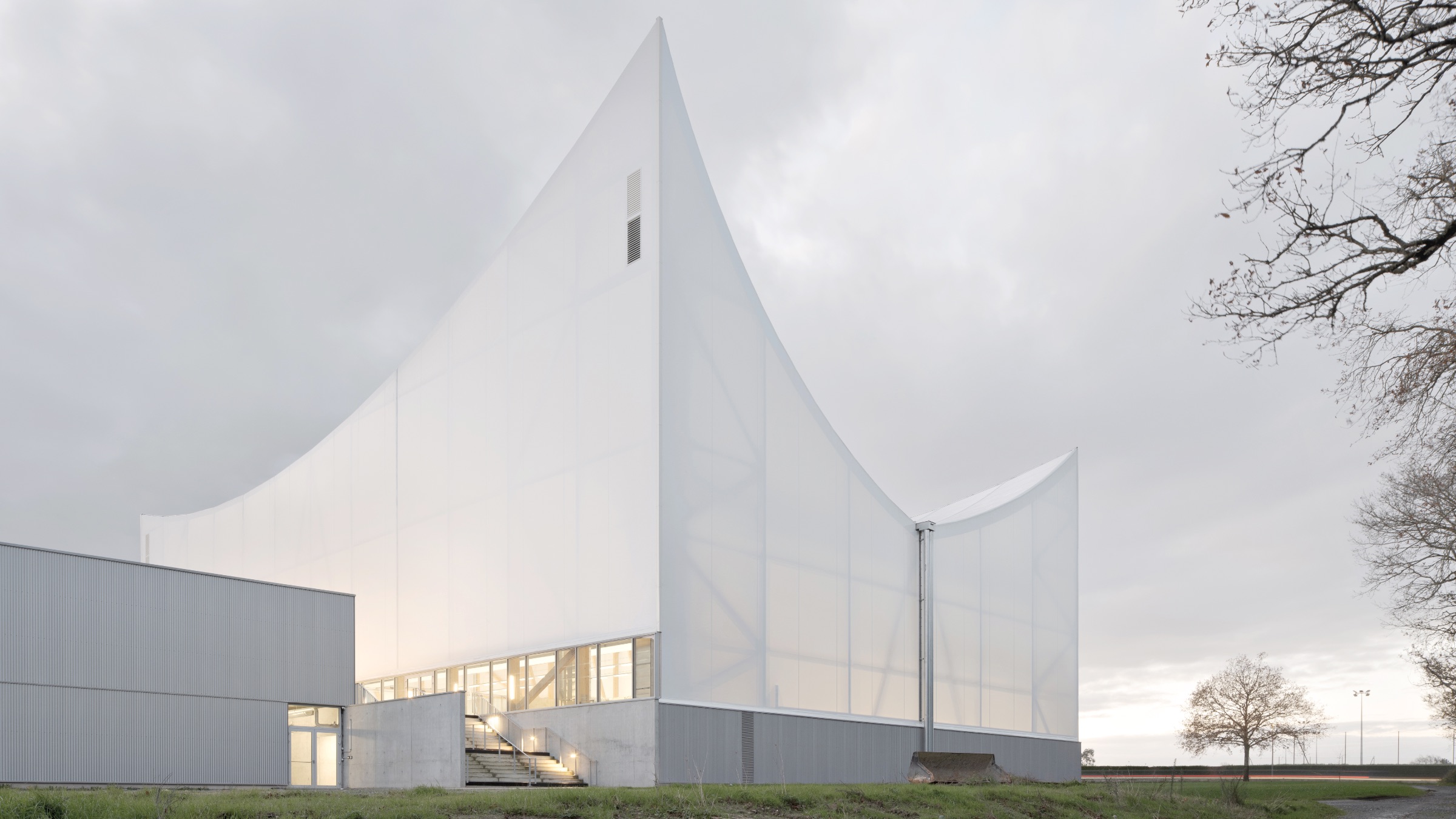 A Civic Canopy in Rural France by Onze04: A modern white building with sharp, angular rooflines and translucent walls stands on a grassy area under a cloudy sky.
