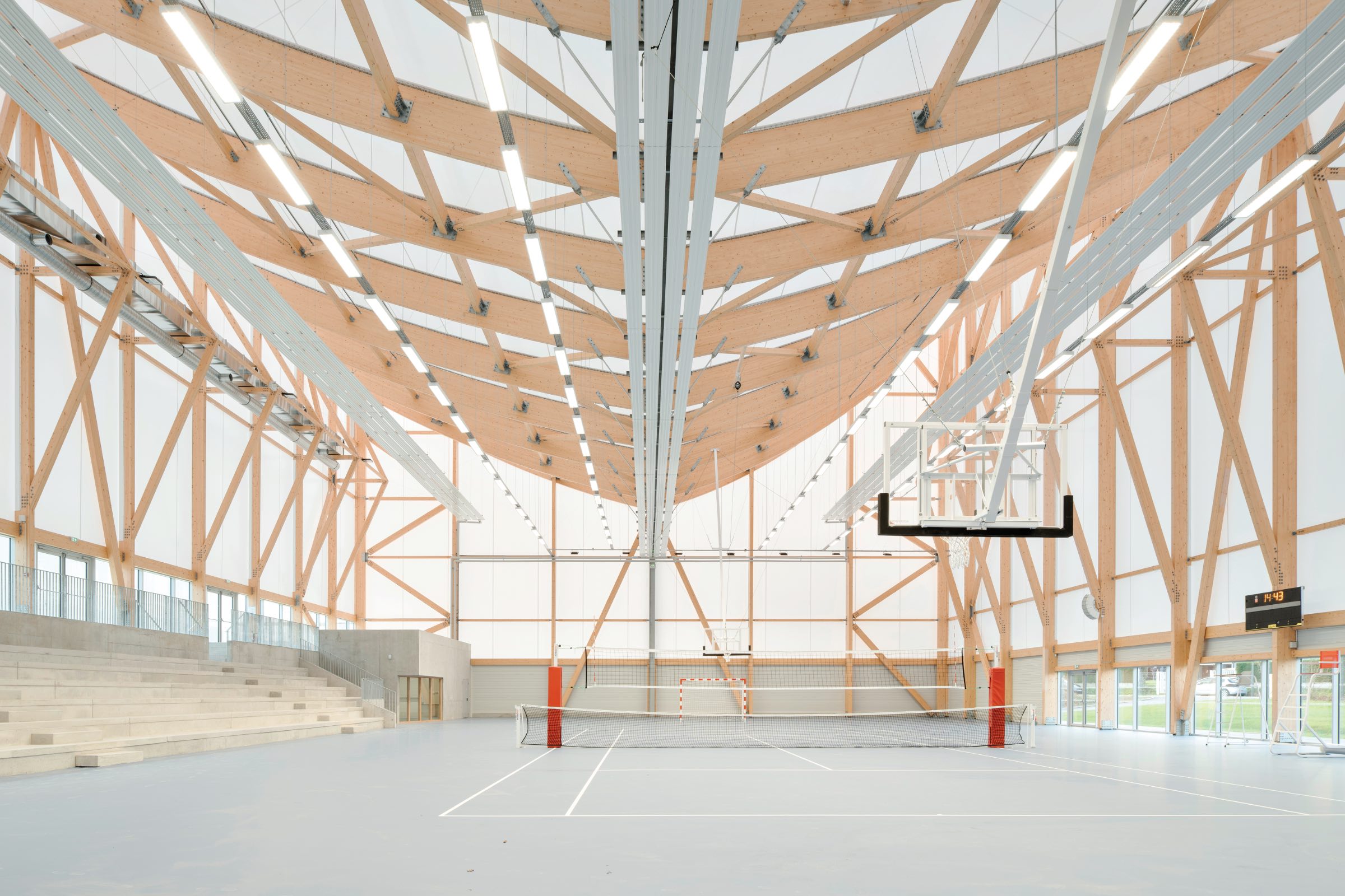 Interior of a modern gymnasium by Onze04 with a wooden truss ceiling, volleyball net, basketball hoop, bleachers on the left, and large windows allowing natural light.