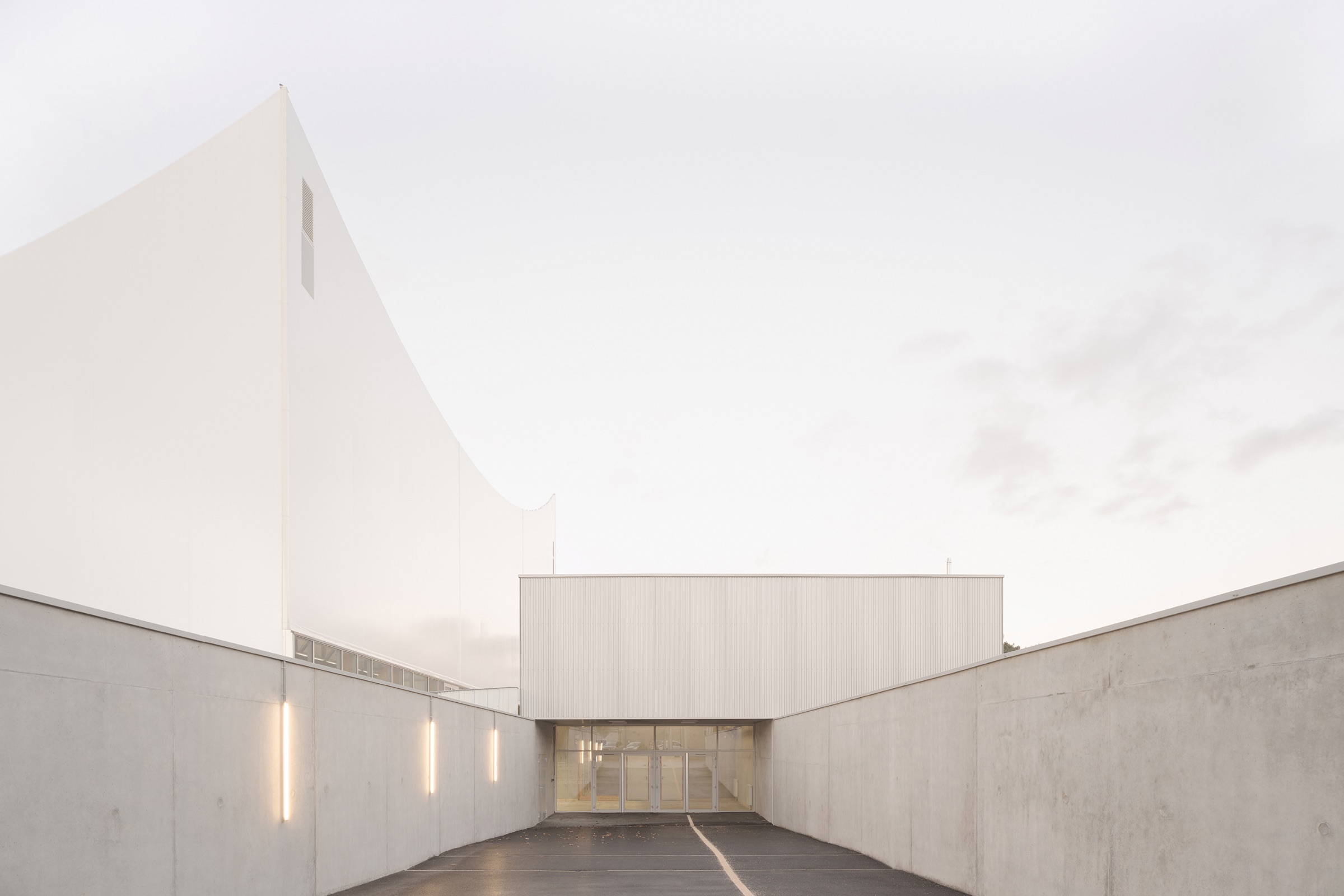 Minimalist white building by Onze04 with geometric shapes, viewed from a concrete driveway, under a pale sky. Vertical lights are mounted on both side walls near the entrance.