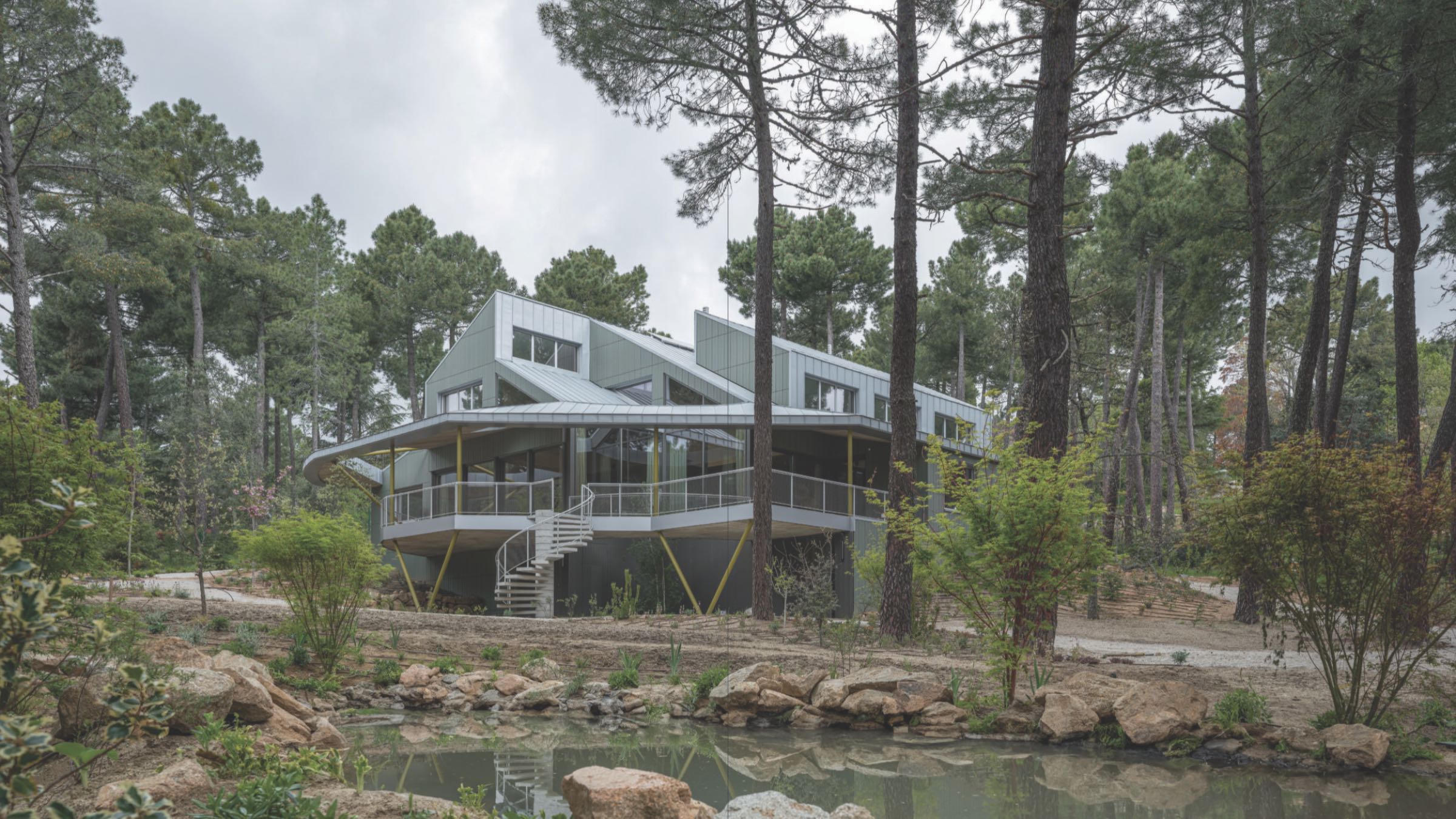 Modern two-story house with angular roof and large windows, elevated on stilts, surrounded by trees and plants, with a small pond in the foreground—an embodiment of Antifascist Architecture that emphasizes openness, harmony with nature, and rejection of oppressive designs.