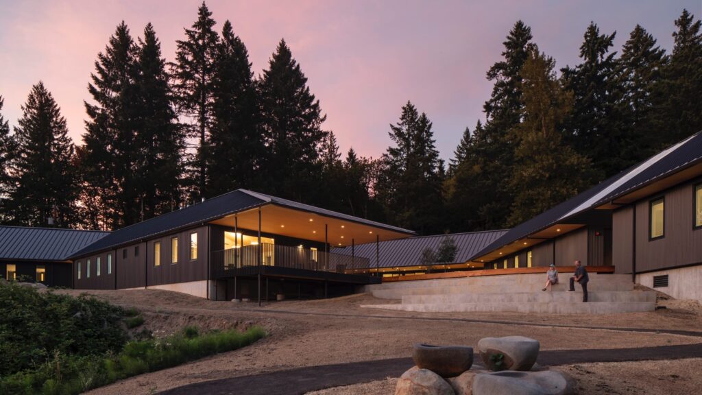 Modern, single-story building with large windows, surrounded by tall trees at sunset. Two people sit on outdoor steps near a landscaped area with large rocks.