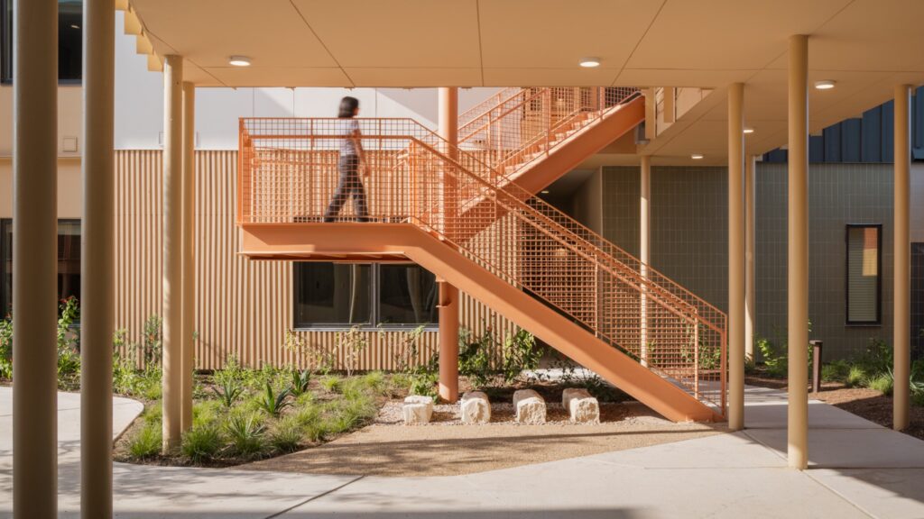 A person walks up an orange outdoor staircase attached to a modern building with a landscaped area below.