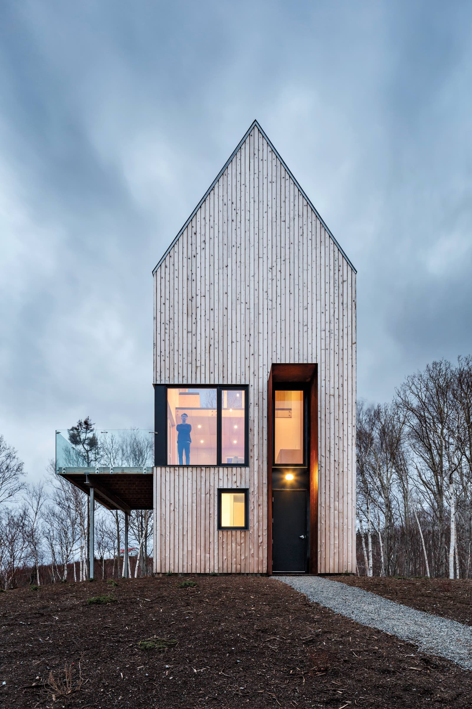 A modern, narrow wooden house by Omar Gandhi with large windows, a glass balcony, and a person visible inside, set against a cloudy sky and surrounded by leafless trees.