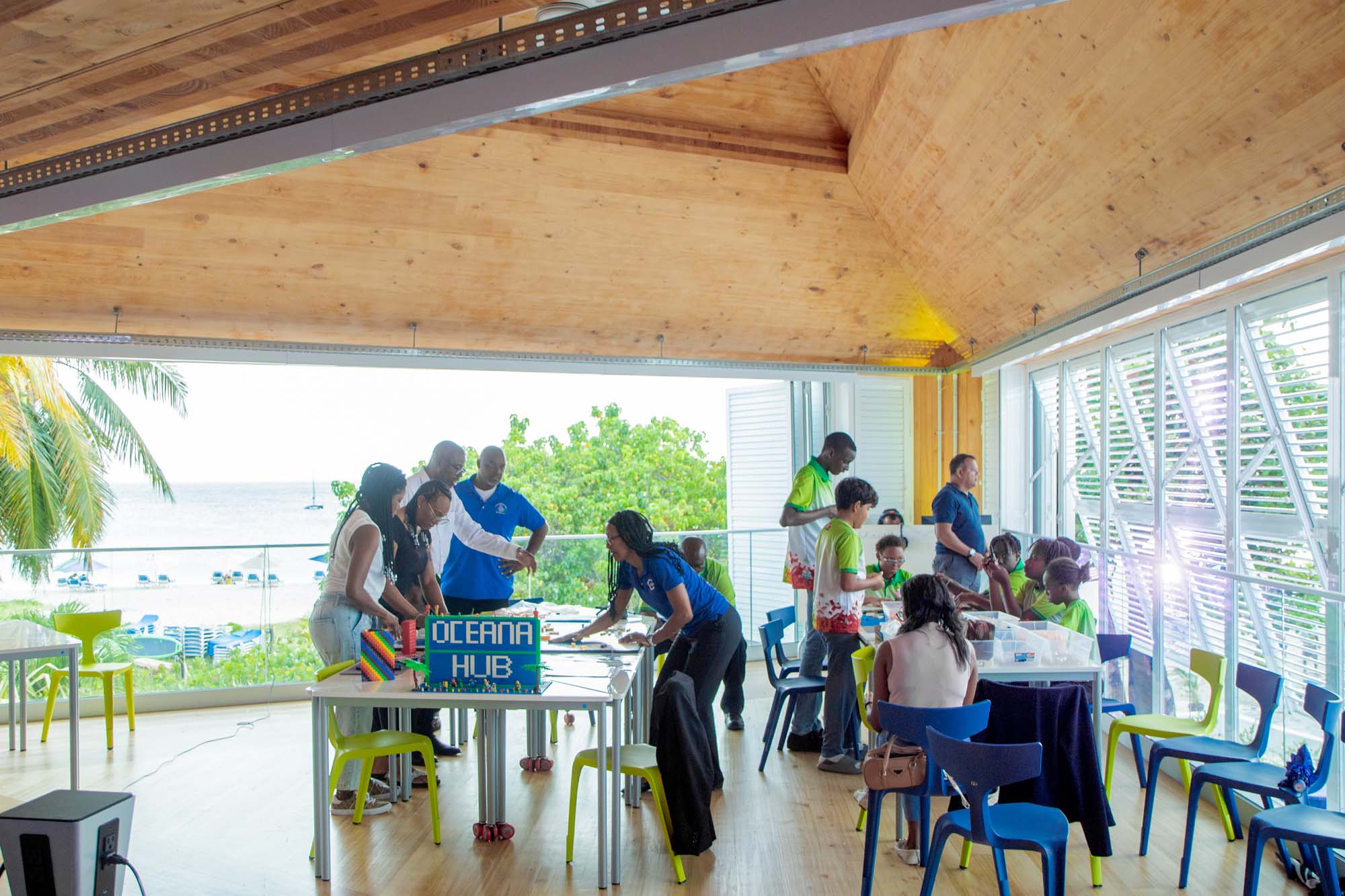 People gather in a bright room with large windows and a view of the sea, participating in activities around tables labeled “Oceana Hub.”.