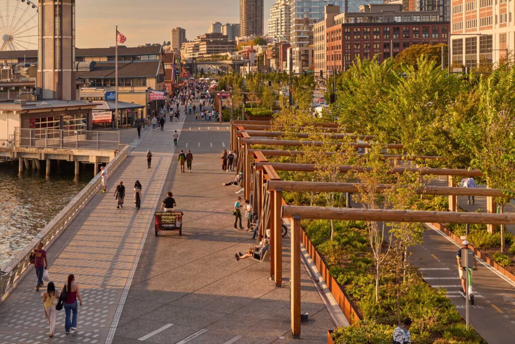 People walk and relax along a waterfront promenade with greenery, wooden pergolas, and city buildings in the background during sunset.