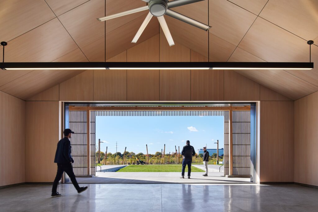 A modern wooden pavilion with an open wall, ceiling fan, and people walking inside and outside, overlooking a grassy area and blue sky.