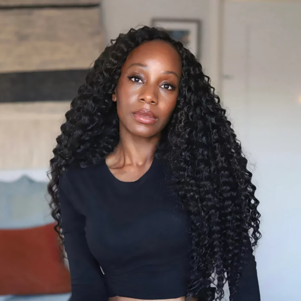 Woman with long, curly black hair wearing a fitted black long-sleeve top, standing indoors with a neutral expression, as if preparing to attend the Trailblazer Awards.