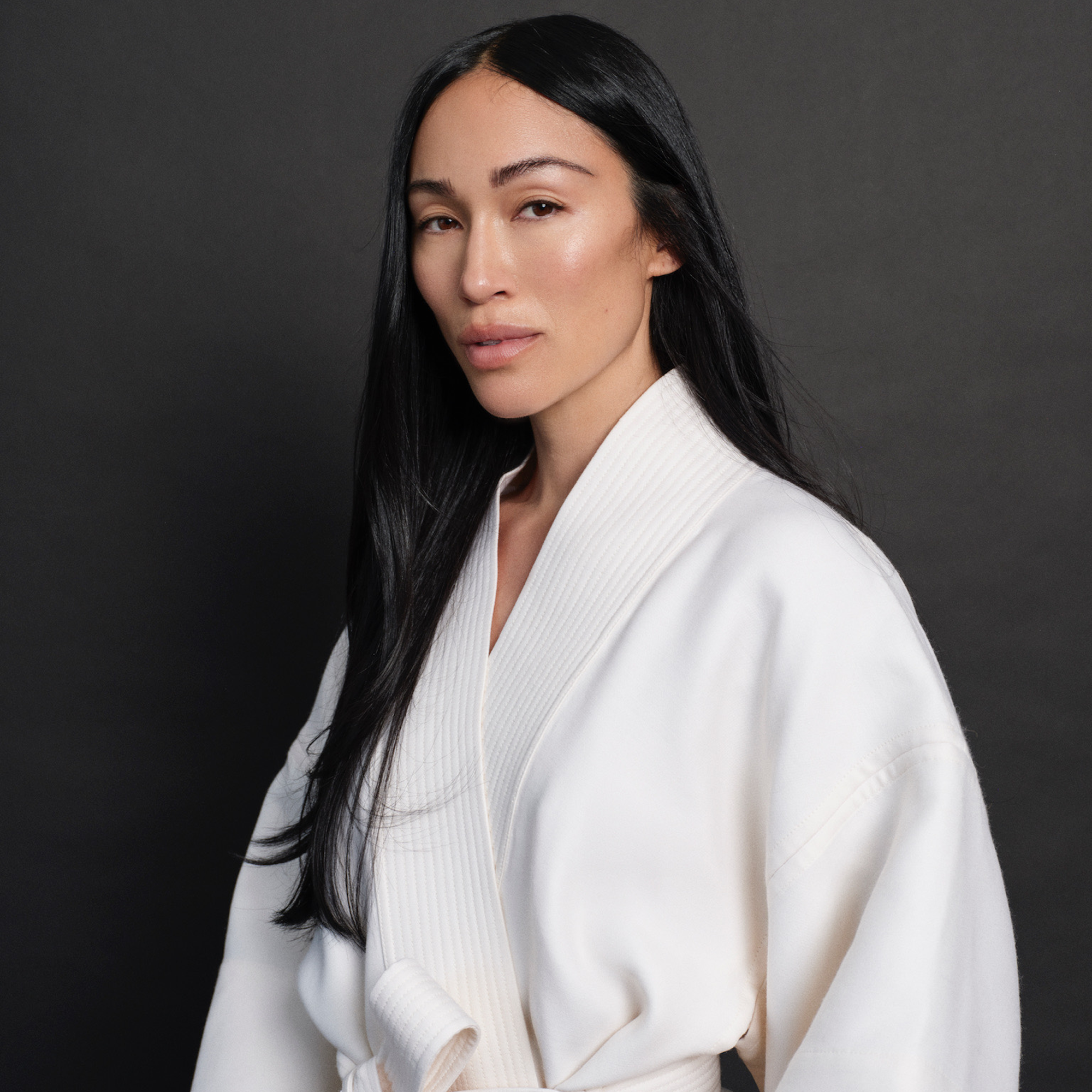 A person with long black hair wearing a white robe stands in front of a dark background, looking at the camera with a neutral expression, reminiscent of an honoree at the Trailblazer Awards.