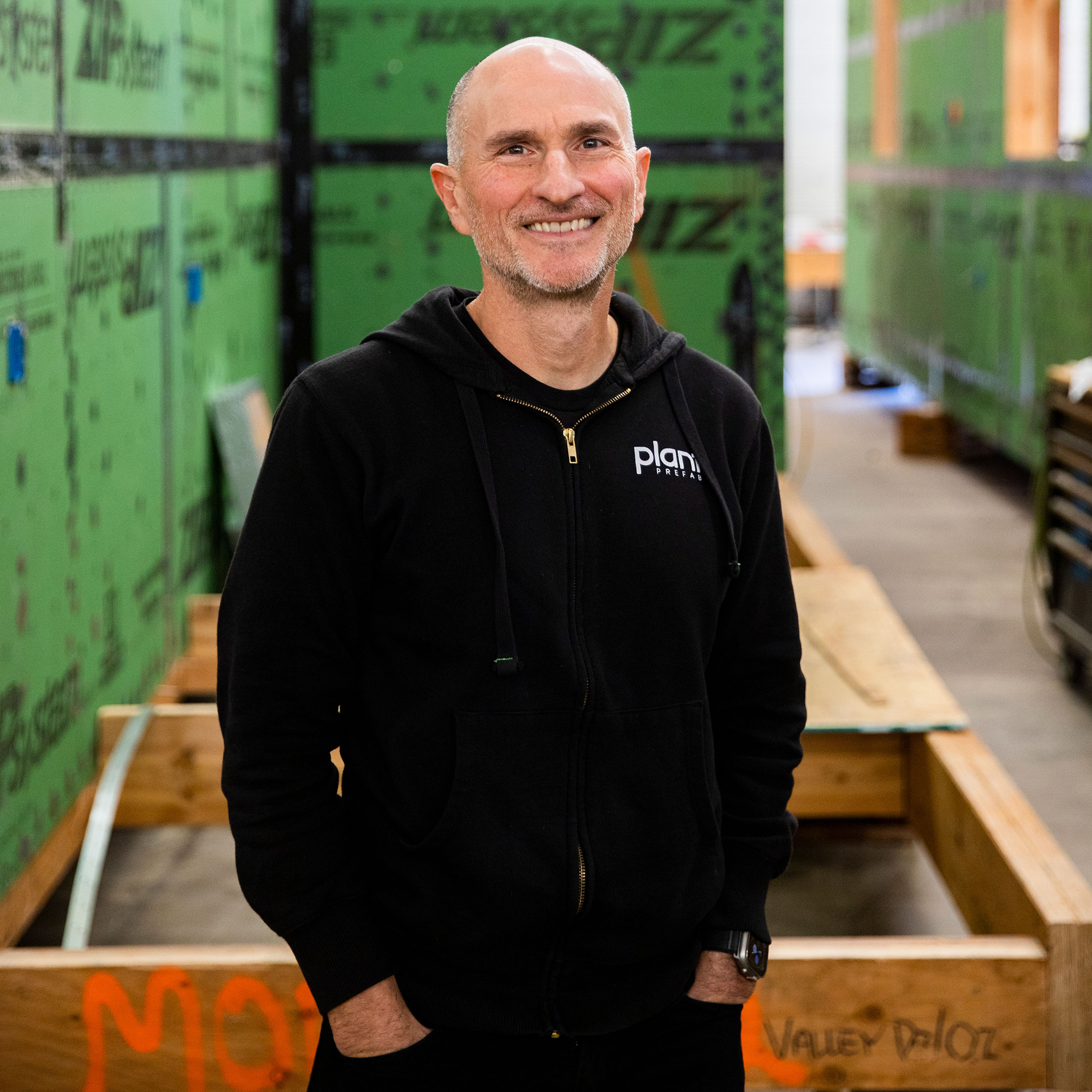 A man in a black hoodie stands and smiles in a warehouse with green construction panels and wooden framing in the background, embodying the innovation celebrated by the Trailblazer Awards.