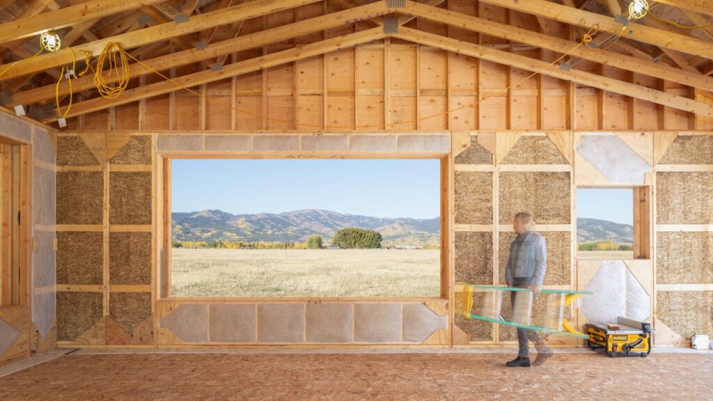 Person carrying a sheet of glass inside a wooden-framed building under construction, with a large window opening showing a scenic mountain landscape outside.