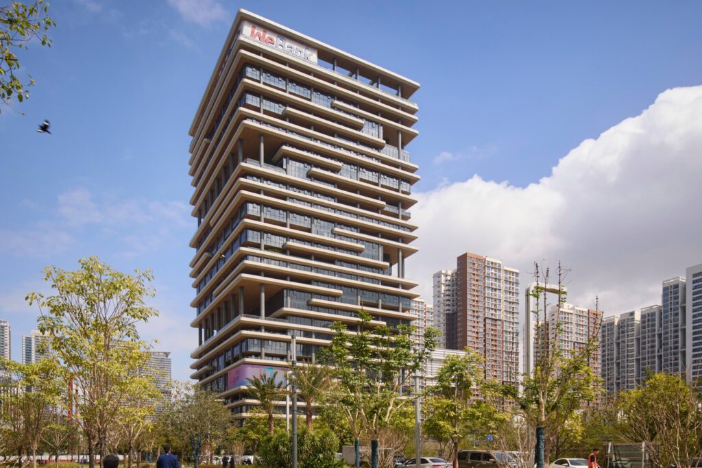 Modern multi-story office building with staggered balconies, surrounded by trees and other high-rise structures, under a blue sky with some clouds.